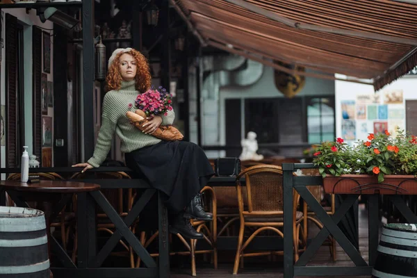 Μια γυναίκα σε ένα μπερέ και ένα πράσινο πουλόβερ κρατά ένα μπουκέτο λουλούδια σε ένα εστιατόριο φόντο — Φωτογραφία Αρχείου