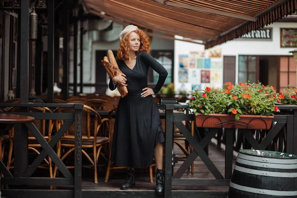 Μια γυναίκα σε ένα μπερέ και ένα μαύρο φόρεμα κρατά μια μπαγκέτα στα χέρια της το c φόντο — Φωτογραφία Αρχείου