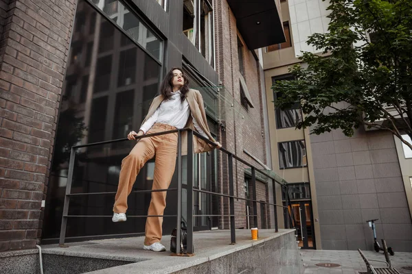 Alta chica con estilo con chaqueta de gran tamaño posando cerca de la barandilla y los pasos en el fondo de la ciudad — Foto de Stock