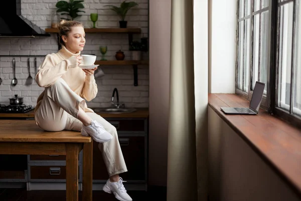 행복 한 젊은 여성이 아늑 한 가정 분위기 속에서 식탁에 앉아 손에 커피를 들고 있다 — 스톡 사진