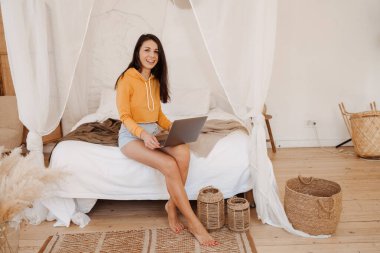 Yatakta dizüstü bilgisayar kullanan ve yatak odasında çalışan serbest çalışan genç bir kadın. 