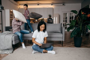 Mutlu Afrikalı genç anne dizüstü bilgisayarla çalışıyor ve kanepede yastıklarla savaşan gürültücü çocuklara adapte oluyor.