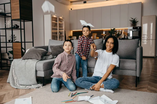 Feliz familia afroamericana, madre y dos hijos que se divierten en casa y vuelan aviones de papel —  Fotos de Stock