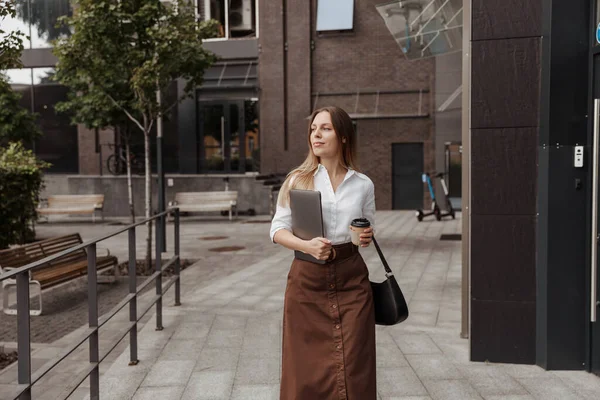 Joven mujer de negocios sosteniendo café y portátil del moderno fondo del edificio de oficinas — Foto de Stock