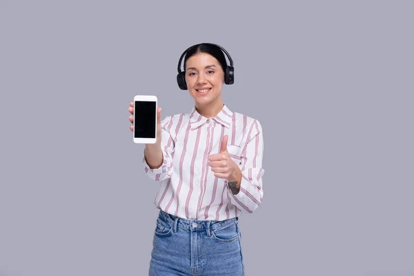 Chica Mostrando Teléfono Usando Auriculares De Pie Aislado con Pulgar hacia Arriba. Commecial, Compras, Concepto de Publicidad Imagen de archivo