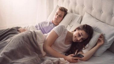 Telefonları kullanarak yatakta yatan bir çift. Kıskanç adam. Güneşli bir sabahta yatakta bir çift.