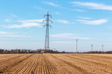 Alanlar arasında duran yüksek voltajlı elektrik direği. Arka planda mavi gökyüzü.