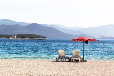 Beautiful pebble beach and there are two white sunbeds with a red parasol. In the background the blue sea and mountains.
