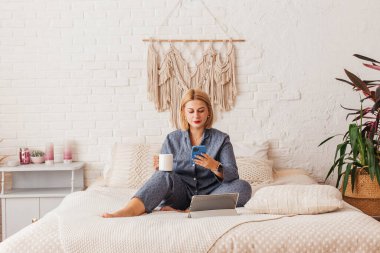 Pijamalı, yatakta dizüstü bilgisayarla çalışan, telefonda konuşan güzel bir genç kadın. İnternetten alışveriş