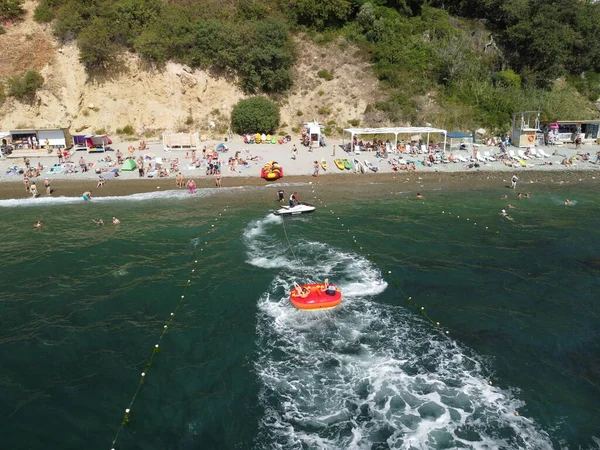 Happy people swim on air mattress behind a high speed water bike. Tourists ride on inflatable watercraft boat. Sea attraction. Water boat skating people on large Inflatable raft. Aerial copter view