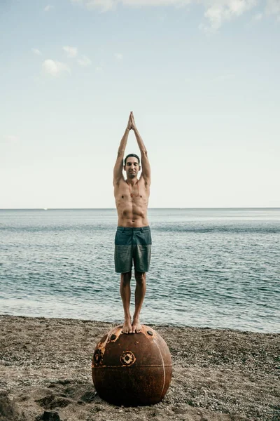 Man doing yoga poses standing on old rusty floating marine mine on the beach with rocky shore and sea background