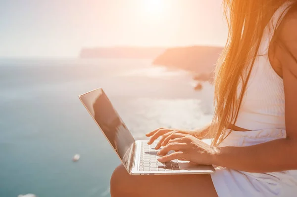 Successful business woman in yellow hat working on laptop by the sea. Pretty lady typing on computer at summer day outdoors. Freelance, travel and holidays concept.