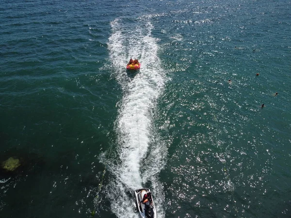 Happy people swim on air mattress behind a high speed water bike. Tourists ride on inflatable watercraft boat. Sea attraction. Water boat skating people on large Inflatable raft. Aerial copter view