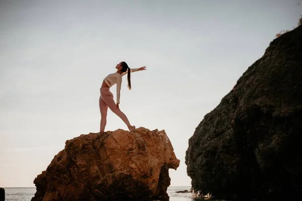 Young woman with black hair, fitness instructor in pink sports leggings and tops, doing pilates on yoga mat with magic pilates ring by the sea on the beach. Female fitness daily yoga concept