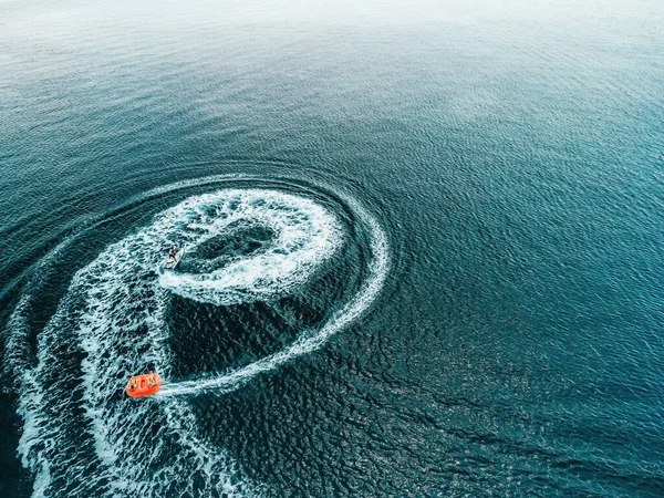 Happy people swim on air mattress behind a high speed water bike. Tourists ride on inflatable watercraft boat. Sea attraction. Water boat skating people on large Inflatable raft. Aerial copter view