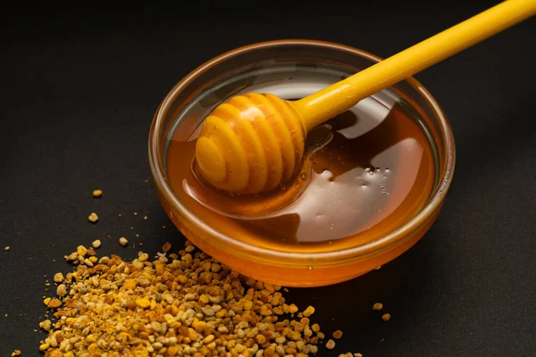 Un cucchiaio giallo di miele immerso in un barattolo di miele e pane d'api sparsi su uno sfondo nero. Miele gocciolante intorno, foto belle e invitanti. Concetto di cibo sano. — Foto Stock