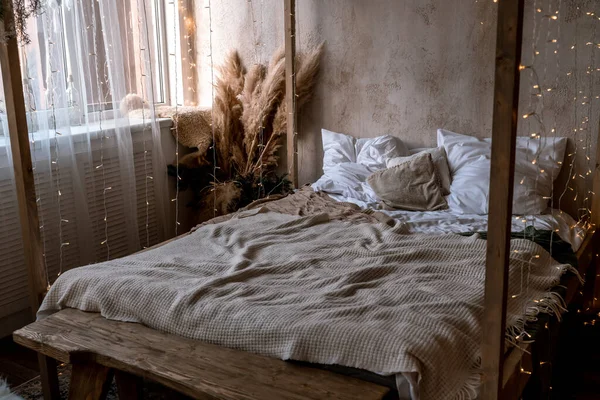 Home interior with four poster bed. There are many white and beige pillows and bedspreads on the bed. A bouquet of dried flowers stands nearby. The bed is decorated with a garland.