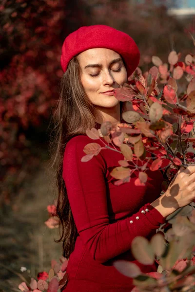 Jeune belle femme dans un pull rouge et béret posant dans la forêt d'automne, sur un fond de feuillage rouge. — Photo