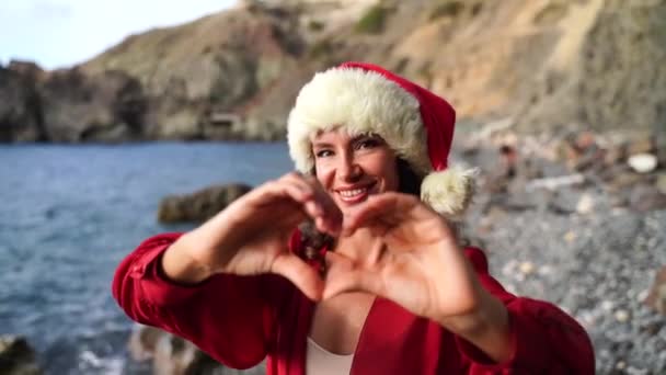 Portrait d'une femme maquillée souriant à la caméra et jouant dans un chapeau de Père Noël sur la plage, au bord de la mer. 4K — Video