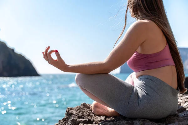 En ung kvinna i grå leggings och en rosa topp med långt löst hår gör yoga utomhus vid havet en solig dag. Kvinnors yoga, kondition, pilates. Begreppet en hälsosam livsstil, harmoni. — Stockfoto