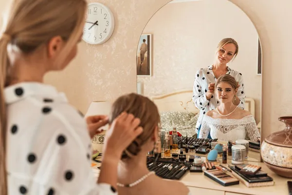 A maquilhadora faz maquiagem profissional para uma jovem, uma noiva. A noiva em um vestido branco, um colar de pérolas e brincos. Artista de maquiagem pinta os lábios das noivas. — Fotografia de Stock