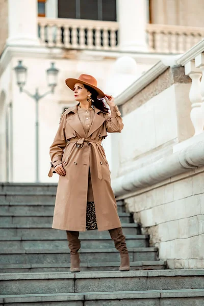 Retrato de moda al aire libre de la joven mujer morena elegante de moda, modelo en sombrero elegante, gargantilla y impermeable ligero posando al atardecer en la ciudad europea. — Foto de Stock