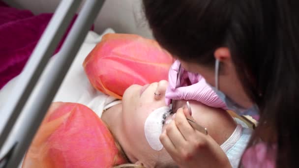 Eyelash extension procedure. Master brunette woman in a pink robe and mask at the workplace in the process of building up. 4K — Stock Video