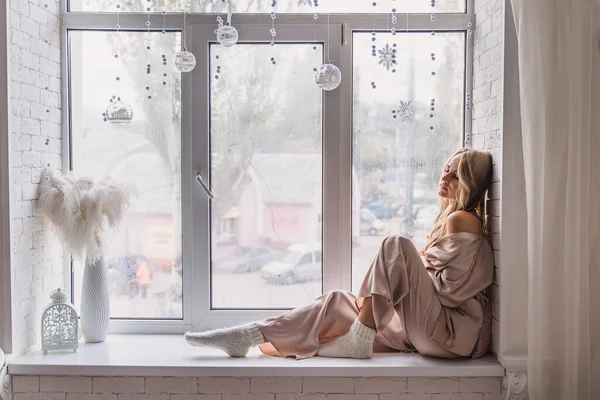 Mujer descansando y pensando en pijamas sentada en el alféizar de la ventana en casa. Vista lateral. —  Fotos de Stock