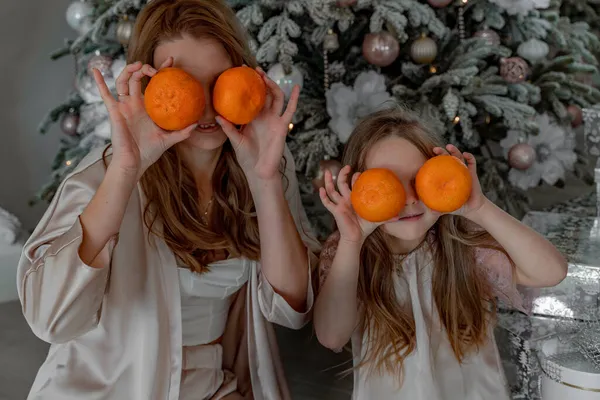 Mamá y su hija sostienen mandarinas a la altura de los ojos en lugar de anteojos. Están sentados bajo un árbol de Navidad decorado en pijama. El concepto de Navidad, vacaciones en familia —  Fotos de Stock