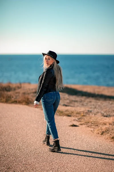 Blondýnka ve stylové černé kožené bundě kráčí podél pobřeží. — Stock fotografie