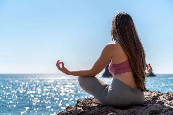 A young woman in gray leggings and a pink top with long loose hair does yoga outdoors by the sea on a sunny day. Womens yoga, fitness, Pilates. The concept of a healthy lifestyle, harmony. — Stock Photo, Image