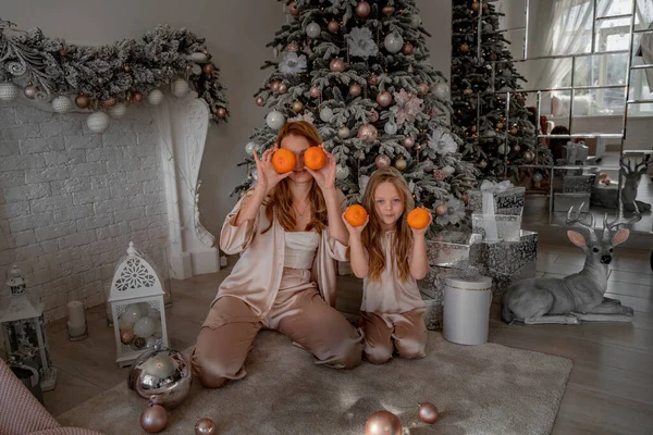 Mamá y su hija sostienen mandarinas a la altura de los ojos en lugar de anteojos. Están sentados bajo un árbol de Navidad decorado en pijama. El concepto de Navidad, vacaciones en familia —  Fotos de Stock