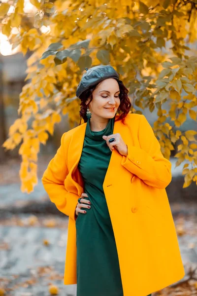 Hermosa mujer camina al aire libre en otoño. Lleva un abrigo amarillo y un vestido verde. Mujer joven disfrutando del clima otoñal. Contenido de otoño. —  Fotos de Stock