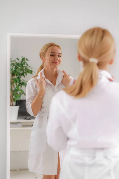 Uma mulher loira com roupas formais brancas olha para si mesma no espelho. Em um fundo branco, folhas verdes na parte de trás. — Fotografia de Stock