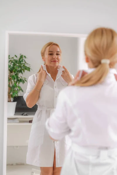 Uma mulher loira com roupas formais brancas olha para si mesma no espelho. Em um fundo branco, folhas verdes na parte de trás. — Fotografia de Stock