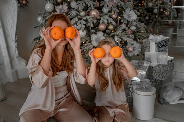 母親と娘はメガネの代わりに目のレベルでみかんを保持しています。彼らはパジャマで装飾されたクリスマスツリーの下に座っています。クリスマス、家族の休日の概念 — ストック写真