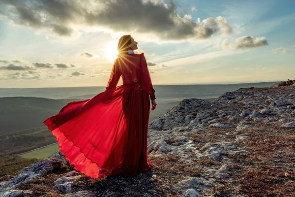 Gizemlerin yükselişi. Bulutların üzerinde uzun kırmızı elbiseli bir kızla gün batımı. Büyülü bir manzarayla dağda duruyor. — Stok fotoğraf