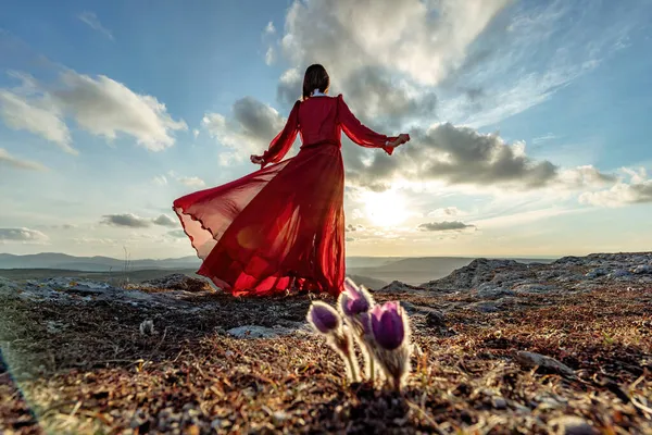 El ascenso de la mística. puesta de sol sobre las nubes con una chica en un vestido rojo largo. Hay una hierba de ensueño en el prado con flores púrpuras. —  Fotos de Stock