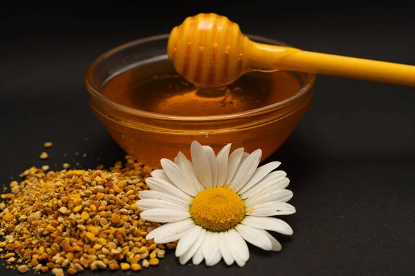 Un cucchiaio giallo di miele immerso in un barattolo di miele e pane d'api sparsi su uno sfondo nero decorato con camomilla viva. Miele gocciolante intorno, foto belle e invitanti. Concetto di cibo sano. — Foto Stock