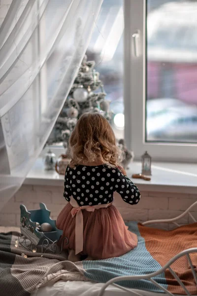 A little girl is sitting on the bed by the window and decorating a small tree with tiny Christmas toys. Happy healthy child celebrating a traditional family holiday. Adorable baby. — Stock Photo, Image