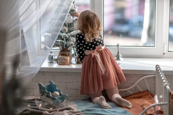 Маленькая девочка сидит на кровати у окна и украшает маленькую елку крошечными рождественскими игрушками. Счастливого здорового ребенка, отмечающего традиционный семейный праздник. Очаровательный ребенок. — стоковое фото