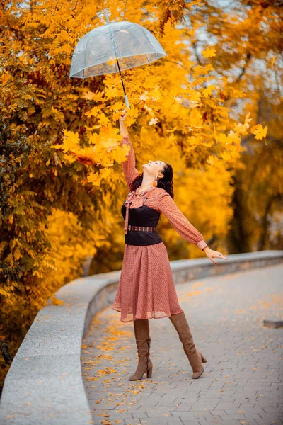 Hermosa chica en un vestido con un paraguas en el parque de otoño. Ella lo sostiene sobre su cabeza, las hojas de otoño caen de él —  Fotos de Stock