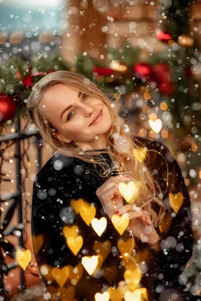Joven rubia con el pelo largo cerca de la cafetería, decorada para Navidad. Ella está vestida con un abrigo de piel corta negro y un vestido corto azul. —  Fotos de Stock
