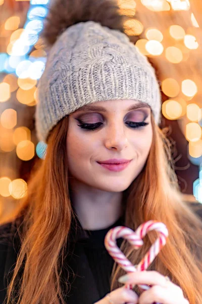 Ένα νεαρό κορίτσι με μακριά μαλλιά σε ένα γκρι καπέλο και με φωτεινό μακιγιάζ κοιτάζει μια καρδιά από ζαχαροκάλαμο στα χέρια της. Στο φόντο των Χριστουγέννων bokeh — Φωτογραφία Αρχείου