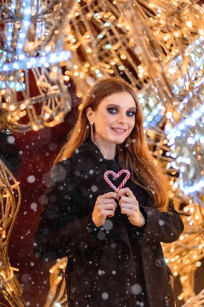 Ένα νεαρό κορίτσι με μακριά μαλλιά με φωτεινό μακιγιάζ σε μαύρο παλτό κοιτάζει την κάμερα. Κρατάει μια καρδιά φτιαγμένη από ζαχαρωτά. Στο φόντο των Χριστουγέννων bokeh — Φωτογραφία Αρχείου