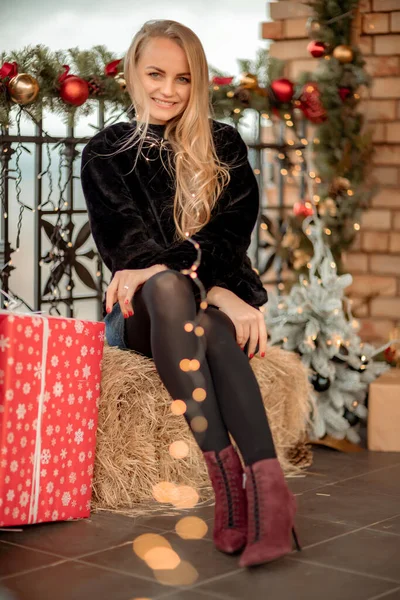 Joven rubia con el pelo largo cerca de la cafetería, decorada para Navidad. Ella está vestida con un abrigo de piel corta negro y un vestido corto azul. —  Fotos de Stock