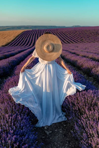 Encantadora mujer joven con un sombrero y vestido blanco en un campo de lavanda púrpura. Estilo de vida al aire libre. Vista trasera —  Fotos de Stock