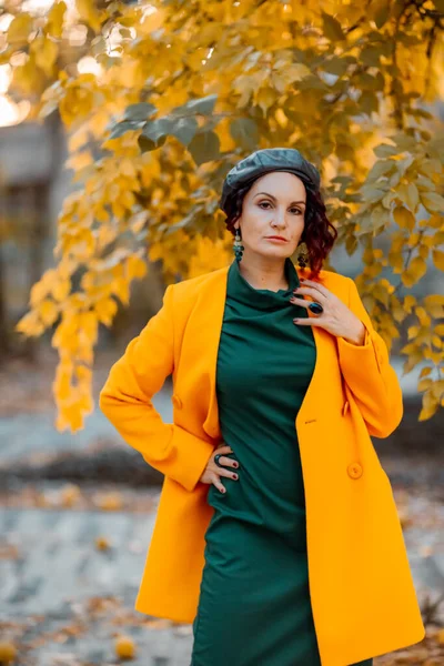 Piękna kobieta wychodzi jesienią na zewnątrz. Ma na sobie żółty płaszcz i zieloną sukienkę. Młoda kobieta ciesząca się jesienną pogodą. Zawartość jesieni. — Zdjęcie stockowe
