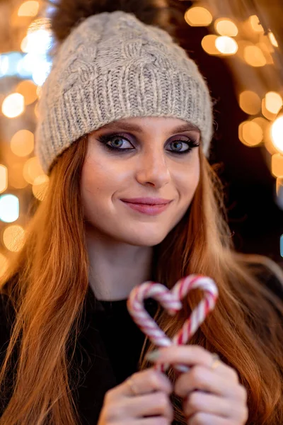 Ένα νεαρό κορίτσι με μακριά μαλλιά σε ένα γκρι καπέλο και με φωτεινό μακιγιάζ κοιτάζει την κάμερα. Κρατάει μια καρδιά φτιαγμένη από ζαχαρωτά. Στο φόντο των Χριστουγέννων bokeh — Φωτογραφία Αρχείου