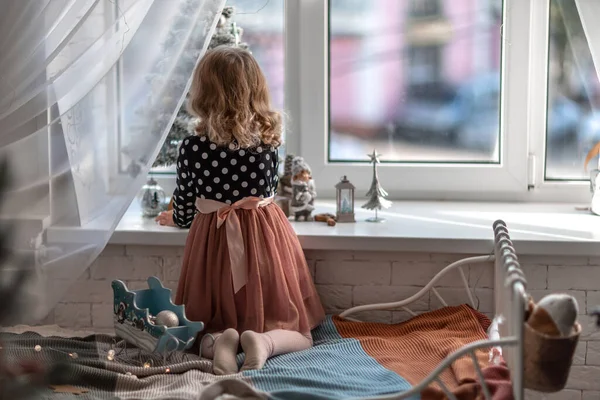 Una niña está sentada en la cama junto a la ventana y decorando un pequeño árbol con pequeños juguetes de Navidad. Feliz niño sano celebrando una fiesta familiar tradicional. Adorable bebé.. — Foto de Stock